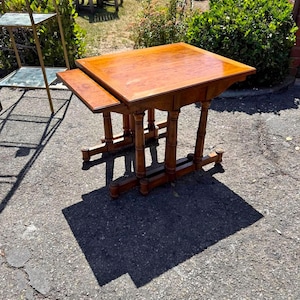 Può includere: Un set di tavolini a incastro in legno marrone medio con piano rettangolare dalla finitura liscia. Il tavolo poggia su quattro gambe tornite collegate da supporti di base orizzontali, con un tavolino più piccolo parzialmente estratto di lato.
