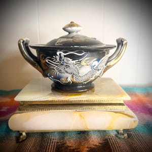 May include: A black ceramic lidded bowl with two handles, featuring a raised silver and blue dragon motif. The bowl rests on a rectangular, polished beige and white stone box with brass trim and small feet, showcasing natural veining patterns.