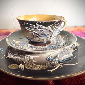 May include: A ceramic tea set consisting of a cup, a saucer, and a plate, all featuring a raised, three-dimensional dragon motif in shades of gray, white, and blue against a dark, matte black background with gold-toned rim accents.