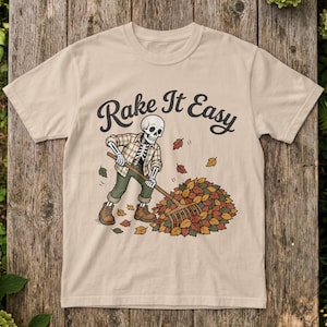 May include: A beige t-shirt featuring a graphic of a skeleton wearing a plaid shirt and green pants, raking a pile of colorful autumn leaves. The text "Rake It Easy" is printed in a black cursive font above the skeleton illustration.