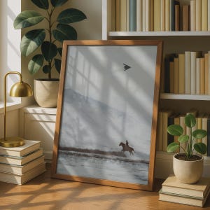 May include: A rectangular wooden frame containing a print of a landscape scene. The artwork depicts a person riding a horse across a snowy, flat terrain with mountains in the background. A small, dark, triangular shape is visible in the sky.