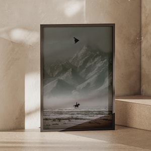 May include: A framed vertical wall art print featuring a monochromatic landscape with a silhouette of a person riding a horse across a flat, misty terrain. In the background, large snow-capped mountains rise under a dark sky with a small black aircraft.