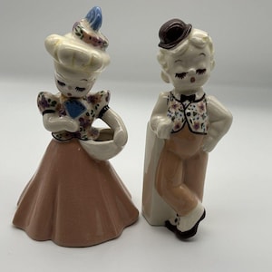 May include: Two ceramic figurines depicting a pair of people. The left figure wears a pink skirt, a floral top, and a blue-accented hat. The right figure wears tan trousers, a floral vest, and a brown bowler hat. Both have painted facial features.