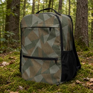May include: A backpack featuring a geometric camouflage pattern in shades of olive green, brown, and tan. The bag includes a top handle, a main zippered compartment, a smaller front zippered pocket, and black mesh side pockets for storage.