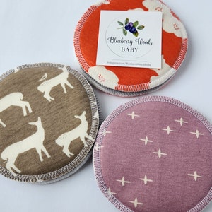 May include: Three round fabric nursing pads with different patterns. The first pad has a brown background with white deer silhouettes. The second pad has an orange background with white clouds and a small blueberry design. The third pad has a purple background with a white cross pattern. The second pad has a white label with the text "Blueberry Woods Baby" and a website address.