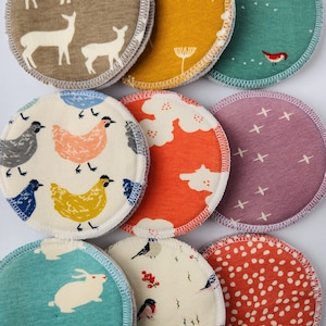 May include: Ten round, fabric nursing pads with various patterns. The patterns include deer, chickens, flowers, cross-stitching, birds, and polka dots. The pads are white with different colored patterns.
