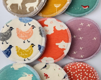 May include: Ten round, fabric nursing pads with various patterns. The patterns include deer, chickens, flowers, cross-stitching, birds, and polka dots. The pads are white with different colored patterns.