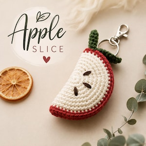 May include: A crocheted apple slice keyring featuring a cream-coloured centre with dark brown embroidered seeds, a red outer edge, and a green stem. The keyring includes a silver-toned metal lobster clasp and ring. Text reads Apple SLICE.