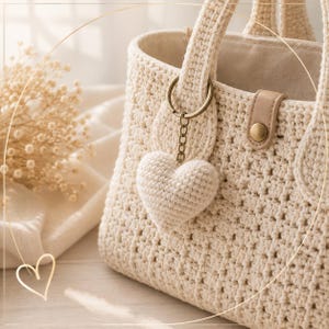 May include: A cream-coloured crocheted handbag featuring a textured stitch pattern, two sturdy handles, and a small beige leather-like tab with a gold-tone snap closure. A matching crocheted heart charm hangs from a gold-tone metal keyring.