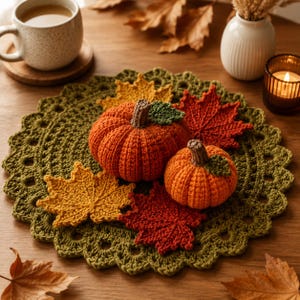 May include: A round olive green crocheted doily featuring two orange crocheted pumpkins with brown stems and green leaves, surrounded by three crocheted maple leaves in shades of yellow, orange, and deep red, arranged on a wooden surface.
