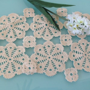 May include: A beige crocheted table runner with a delicate floral pattern. The runner is made of fine yarn and has a soft, airy texture.
