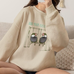 May include: A beige crewneck sweatshirt featuring a graphic illustration of two Java sparrow birds sitting on a swing set. Above the birds, the text reads "ADD YOUR CUSTOM" in green lettering. The sweatshirt has long sleeves and a ribbed neckline.