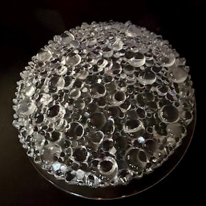 May include: A circular clear glass plate or bowl featuring a textured surface covered in numerous raised, spherical bubble-like protrusions of varying sizes. The design creates a multi-dimensional, refractive effect across the entire top surface.