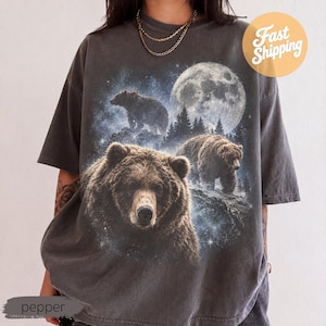 May include: A charcoal grey short-sleeve t-shirt featuring a graphic print of brown bears in a forest setting under a large full moon and starry night sky. The shirt has a relaxed fit and a crew neckline, showcasing a detailed wildlife scene.