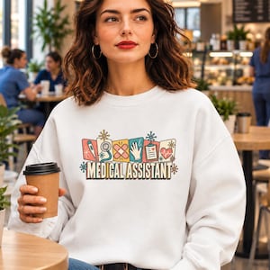 May include: A white long-sleeve sweatshirt featuring a graphic print with the text MEDICAL ASSISTANT. The design includes colourful rectangular icons of a thermometer, stethoscope, bandages, glove, clipboard, and heart, accented with small star shapes.