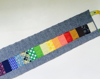 May include: A long, rectangular fabric roll-up organizer featuring a denim-textured gray border. The center panel consists of a vibrant, multi-colored sequence of fabric squares in shades of purple, blue, green, yellow, orange, red, and brown.