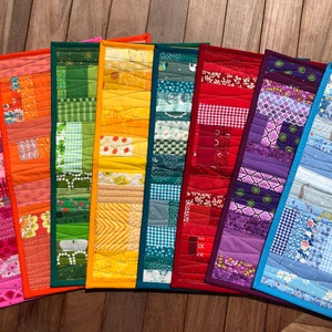 May include: Eight colourful patchwork table runners in a rainbow pattern. The runners are made of different fabrics and have a quilted texture.