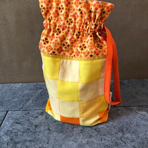 May include: A drawstring bag featuring a top section with a yellow, orange, and white geometric floral pattern. The lower body is constructed from a patchwork of yellow and orange fabric squares. An orange fabric strap is attached to the side.