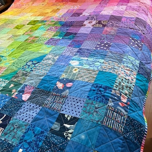 May include: A handmade quilt with a vibrant rainbow gradient design. The quilt features a patchwork pattern of squares in shades of red, orange, yellow, green, blue, and purple. The quilt is made of various fabrics and textures, creating a visually appealing and cozy textile.