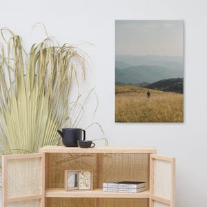 May include: A rectangular wall art print featuring a landscape of a person walking through a golden grassy field with rolling blue mountains in the distance under a pale sky. The scene captures a vast, open outdoor environment with soft lighting.