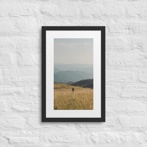 May include: A framed vertical wall art print featuring a landscape photograph of a person walking through a golden grassy field with rolling blue mountains in the distance under a pale sky. The artwork is enclosed in a simple black frame.