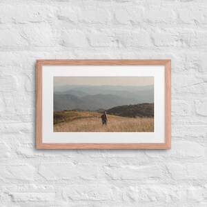 Puede incluir: Un marco de fotos rectangular de madera que contiene una fotografía de paisaje de una persona caminando por un campo de hierba alta y seca hacia cadenas montañosas azules y brumosas bajo un cielo suave, sobre una pared de ladrillo blanco.