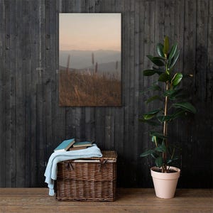 Könnte beinhalten: Ein rechteckiger Wandkunstdruck mit einer Landschaft aus hügeligen Bergen bei Sonnenuntergang, platziert über einem braunen geflochtenen Weidenkorb. Eine hellblaue Strickdecke liegt über dem Korb, in dem sich drei Bücher befinden.