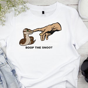May include: White t-shirt with a cartoon snake and a hand reaching out to touch it. The text "BOOP THE SNOOT" is printed below the image.