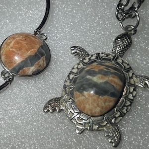 Könnte beinhalten: Ein Schmuckset aus silberfarbenem Metall, bestehend aus einem Armreif und einer Halskette mit schildkrötenförmigem Anhänger. Beide Teile enthalten einen polierten, ovalen Steineinsatz mit wirbelnden Mustern in Beige, Braun und Dunkelgrau.