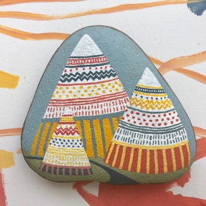 Puede incluir: Pintura de roca pintada a mano que representa tres tipis coloridos con patrones geom&eacute;tricos. Los tipis est&aacute;n pintados en tonos de rojo, amarillo, blanco y negro sobre un fondo gris.