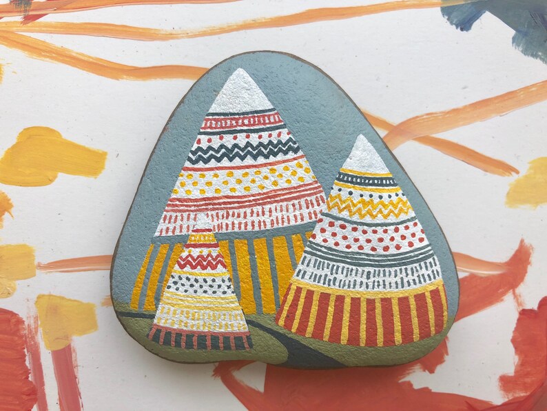 Puede incluir: Pintura de roca pintada a mano que representa tres tipis coloridos con patrones geom&eacute;tricos. Los tipis est&aacute;n pintados en tonos de rojo, amarillo, blanco y negro sobre un fondo gris.