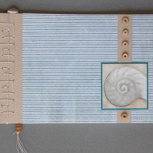 Pode incluir: Diário artesanal com fundo listrado azul e cinza. O diário apresenta um desenho de concha em uma moldura turquesa, botões de madeira e uma lombada costurada decorativa. Uma pequena borla branca pende da parte inferior.