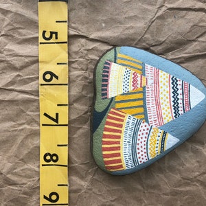 Puede incluir: Piedra pintada a mano con fondo azul y dise&ntilde;os geom&eacute;tricos coloridos. La piedra tiene forma de l&aacute;grima y presenta una variedad de patrones en rojo, amarillo, blanco y negro.
