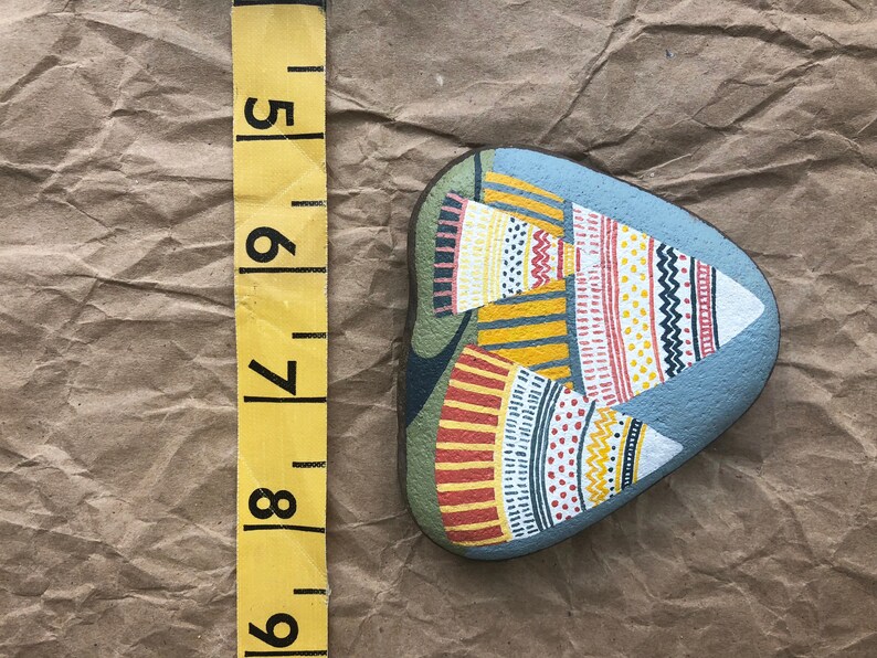 Puede incluir: Piedra pintada a mano con fondo azul y dise&ntilde;os geom&eacute;tricos coloridos. La piedra tiene forma de l&aacute;grima y presenta una variedad de patrones en rojo, amarillo, blanco y negro.