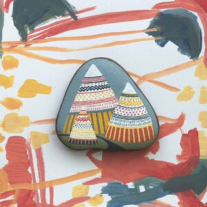 Puede incluir: Una piedra pintada a mano con un fondo gris y dos tipis coloridos. Los tipis est&aacute;n decorados con patrones geom&eacute;tricos rojos, amarillos, blancos y negros.