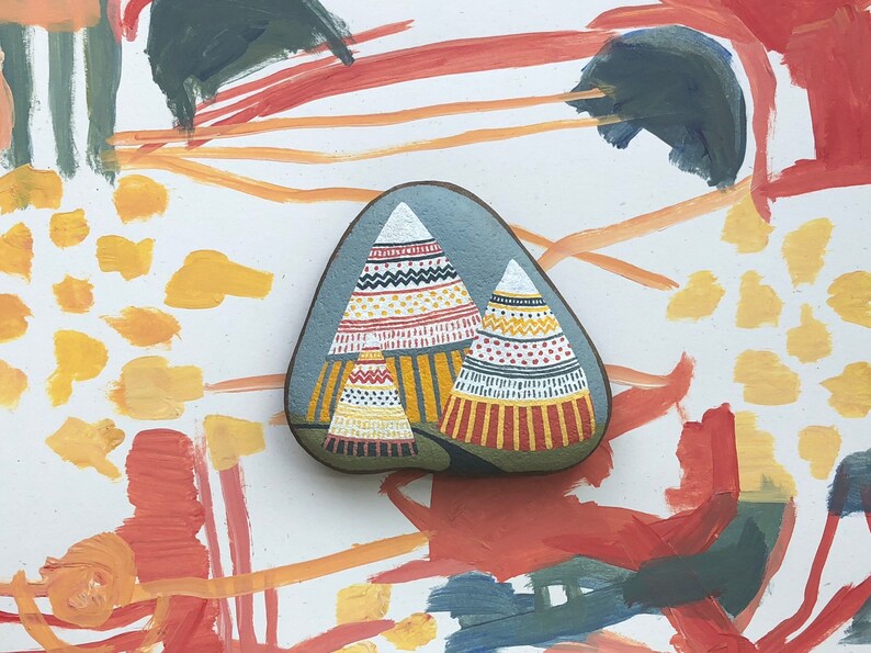 Puede incluir: Una piedra pintada a mano con un fondo gris y dos tipis coloridos. Los tipis est&aacute;n decorados con patrones geom&eacute;tricos rojos, amarillos, blancos y negros.