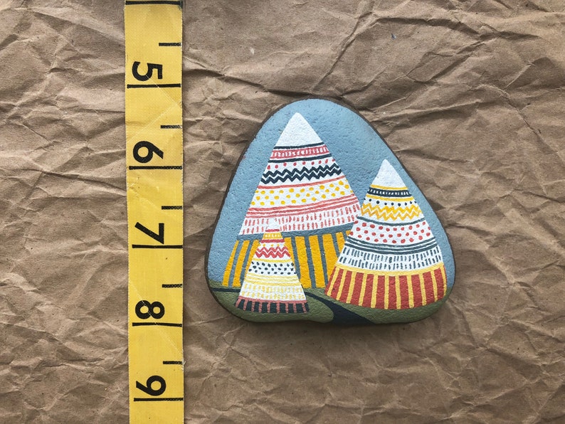 Puede incluir: Pintura de roca pintada a mano que representa tres tipis coloridos con patrones geom&eacute;tricos en tonos rojos, amarillos, blancos y negros. Los tipis est&aacute;n pintados sobre un fondo gris.