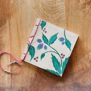 Puede incluir: Un diario cuadrado hecho a mano con un diseño botánico de hojas verdes y bayas moradas. La cubierta está cosida a mano con hilo rojo. Una pulsera de cuentas con cuentas coloridas está unida al diario con una cuerda roja. Sobre una superficie de madera.