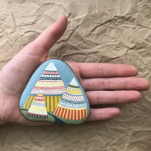 Puede incluir: Piedra pintada a mano con un fondo azul y tres tipis coloridos y estampados. Los tipis son amarillos, rojos y blancos con dise&ntilde;os geom&eacute;tricos.