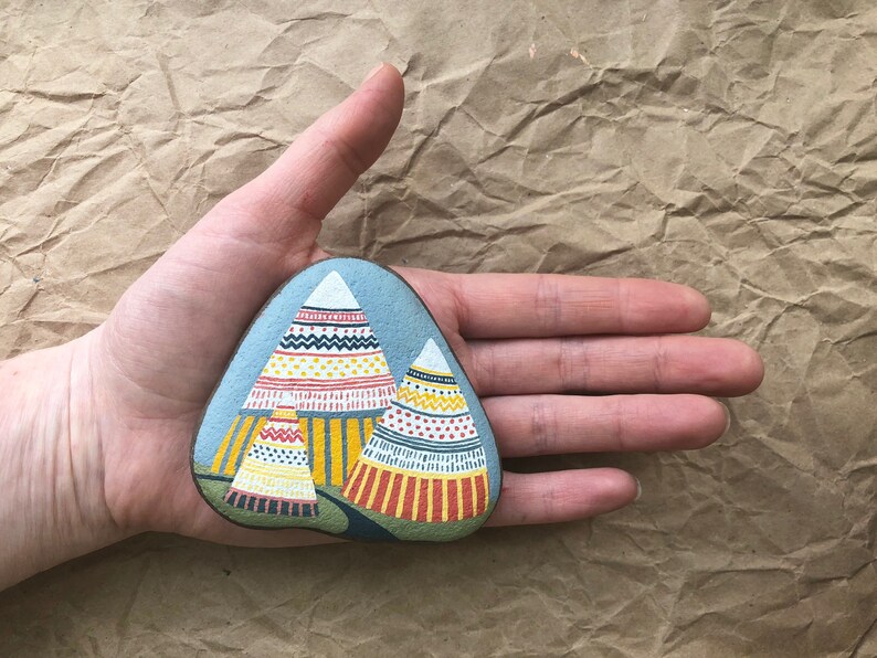 Puede incluir: Piedra pintada a mano con un fondo azul y tres tipis coloridos y estampados. Los tipis son amarillos, rojos y blancos con dise&ntilde;os geom&eacute;tricos.
