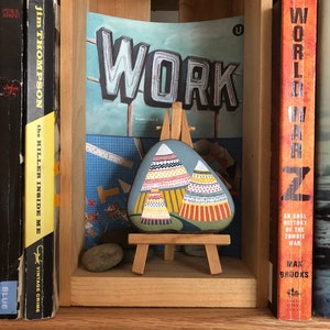 Puede incluir: Una piedra pintada con un dise&ntilde;o de tipis coloridos en un caballete de madera. La piedra est&aacute; en un estante con libros detr&aacute;s. Los libros tienen t&iacute;tulos como "World War Z" y "The Killer Inside Me". Un letrero sobre la piedra dice "WORK".