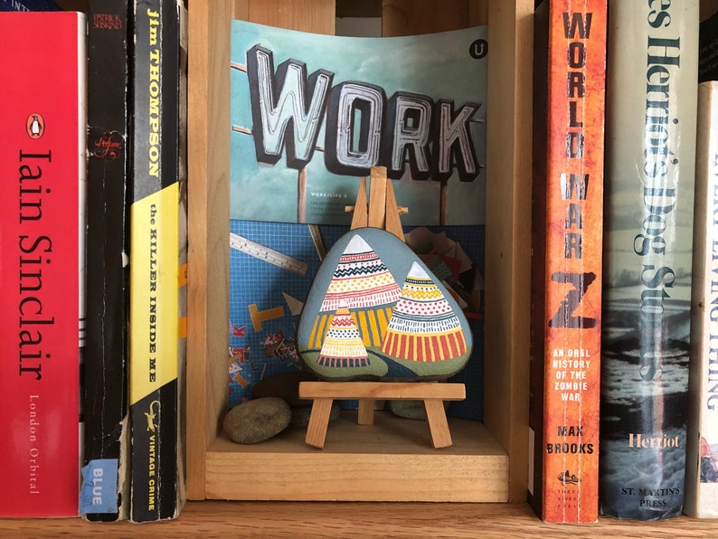 Puede incluir: Una piedra pintada con un dise&ntilde;o de tipis coloridos en un caballete de madera. La piedra est&aacute; en un estante con libros detr&aacute;s. Los libros tienen t&iacute;tulos como "World War Z" y "The Killer Inside Me". Un letrero sobre la piedra dice "WORK".