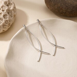 May include: A pair of silver-toned drop earrings featuring two slender, curved metal bars that intersect to form an elongated, open marquise shape. The earrings have a polished, smooth finish and are suspended from small, simple circular loops.