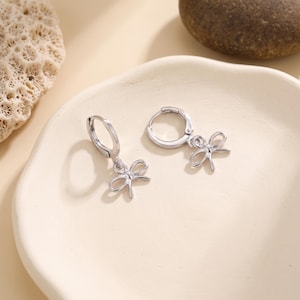 May include: A pair of silver-toned hoop earrings, each featuring a small, delicate dangling charm shaped like a stylized bow. The earrings have a polished metallic finish and a secure hinged clasp mechanism, presenting a clean and simple design.