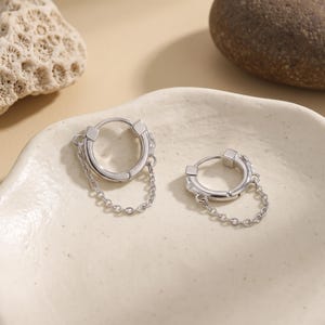 May include: A pair of silver-toned hoop earrings, each featuring a small, polished circular hoop with a delicate, fine-link chain draped across the bottom. The earrings have a smooth, metallic finish and a secure hinged closure mechanism.