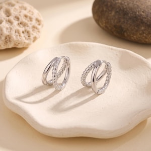 May include: A pair of silver-toned hoop earrings, each featuring a double-band design. One band is smooth and polished, while the other band has a twisted rope texture. These small circular earrings have a hinged closure mechanism.