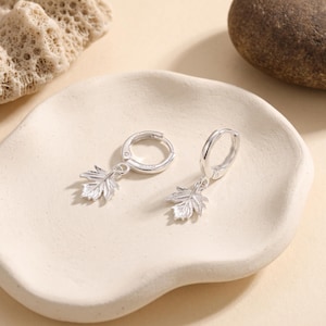May include: A pair of silver hoop earrings featuring dangling maple leaf charms. Each earring consists of a small circular hoop with a textured, detailed leaf pendant attached to the bottom, showcasing a polished metallic finish throughout.
