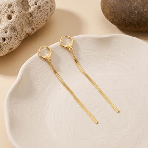 May include: A pair of gold-toned hoop earrings featuring long, slender, flat snake chain dangles. Each earring consists of a small circular hoop closure connected to a flexible, shimmering metallic chain that hangs straight down in a linear design.