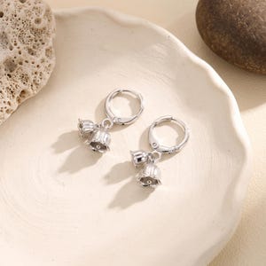 May include: A pair of silver-toned hoop earrings, each featuring two dangling charms shaped like lily of the valley flowers. The earrings have a polished metallic finish and a hinged clasp mechanism, showcasing a detailed floral design.