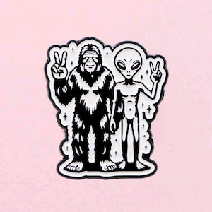 May include: Black and white enamel pin featuring a Bigfoot and an alien, both giving the peace sign. The pin has a white border with star accents and is set against a pink background.